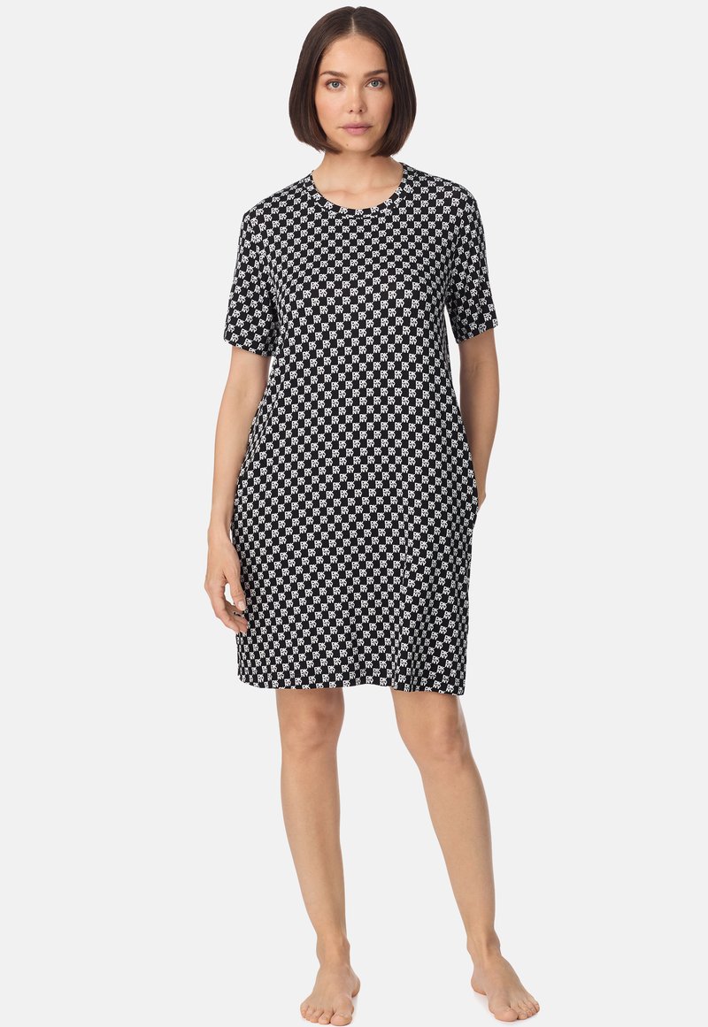 Femme aux cheveux bruns courts portant une robe mi-longue à manches courtes avec un motif noir et blanc, debout pieds nus sur un fond uni.