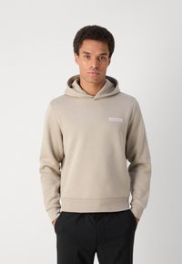 Bež dukserica s kenguru džepom i kapuljačom na vezanje. Ima mali, pravokutni logo Calvin Klein na prsima. Mekana tekstura tkanine.