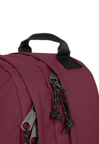 Mörkröd ryggsäck med textilfinish, med svarta dragkedjor, snördrag och ett handtag på toppen för bärande.
