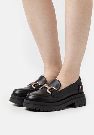 Black leather loafers with a chunky sole, featuring metallic gold hardware accents and a smooth texture. Rounded toe design.