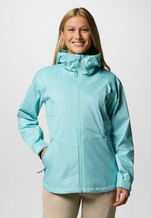 Lächelnde Frau, die eine hellblaue Kapuzenregenjacke und beige Hosen trägt, steht mit einer Hand in der Tasche vor einem einfarbigen Hintergrund.