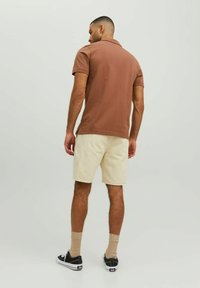 Muž stojaci chrbtom, oblečený v hnedom polo tričku s krátkym rukávom, béžových šortkách, béžových ponožkách a čiernych teniskách na jednolatom svetlom pozadí.