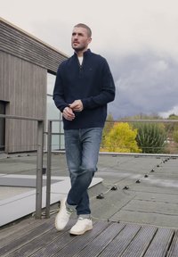 Uomo con maglione blu navy, jeans e sneakers bianche in piedi su un terrazzo sul tetto con ringhiera in legno e cielo nuvoloso sullo sfondo.