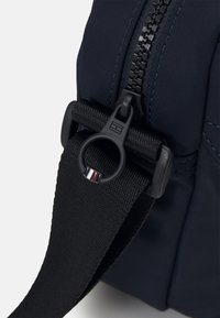 Primo piano della cerniera di una borsa blu marino con un tirante nero e una tracolla nera regolabile attaccata, che presenta un piccolo dettaglio a righe.