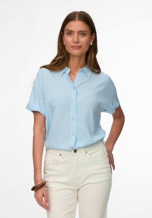 Kobieta z długimi brązowymi włosami, ubrana w jasnoniebieską koszulę na krótki rękaw zapinaną na guziki i białe spodnie, stojąca z jedną ręką w kieszeni.