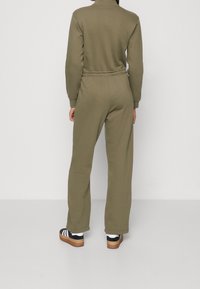 Ensemble de survêtement vert olive à manches longues avec un col montant et une taille élastique. Le bas présente des jambes larges, associé à des baskets noires et blanches.