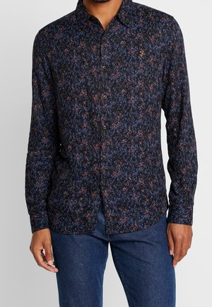 Hombre con una camisa negra de botones con un patrón floral azul y rojo, y jeans azules, posando frente a un fondo gris liso.