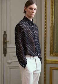 Svart genomskinlig skjorta med knappar och vita prickar, kombinerad med ljusbeige byxor. Har en klassisk krage och figursydda ärmar.