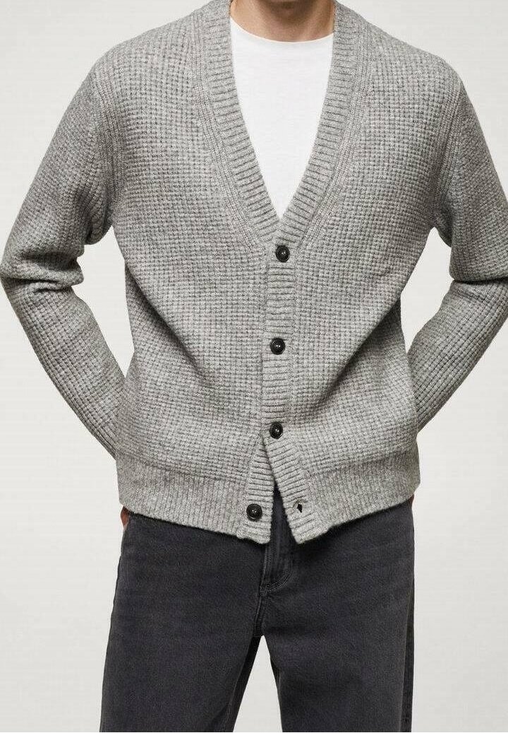 Homme portant un cardigan gris en tricot avec des boutons noirs par-dessus une chemise blanche et un pantalon noir, les bras détendus derrière le dos.