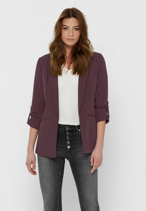 Femme aux longs cheveux bruns portant un blazer violet avec les manches retroussées, un haut blanc et un jean gris taille haute avec des boutons.