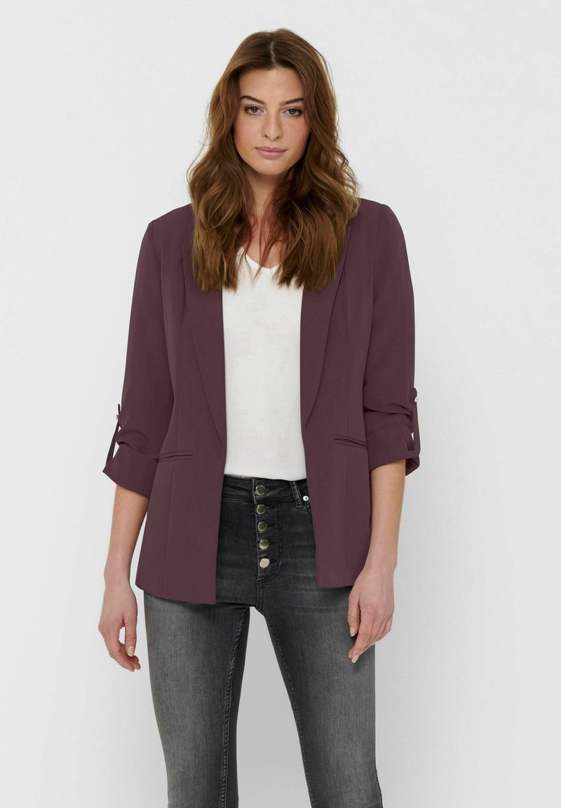 Femme aux longs cheveux bruns portant un blazer violet avec les manches retroussées, un haut blanc et un jean gris taille haute avec des boutons.
