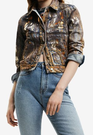 Femme portant une veste en jean courte et délavée multicolore avec des manches retroussées, et un jean taille haute bleu clair.