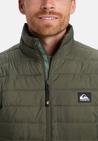 Hombre que lleva una chaqueta acolchada verde oliva con cuello elevado y un parche visible del logo de Quiksilver en el pecho.