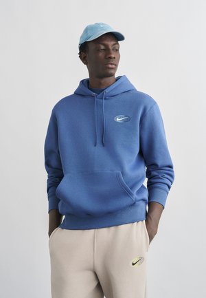 Young man wearing a blue Nike hoodie, beige sweatpants with Nike logo, and a light blue Nike cap, hands in pockets, looking to the side.