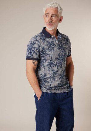 Homme aux cheveux blancs et barbe portant un polo à manches courtes gris et marine avec un imprimé de palmiers, et un pantalon marine, mains dans les poches.