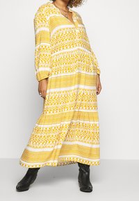 Kvinna klädd i en gul och vit mönstrad lång klänning med puffärmar och svarta ankelboots, stående mot en enfärgad bakgrund.