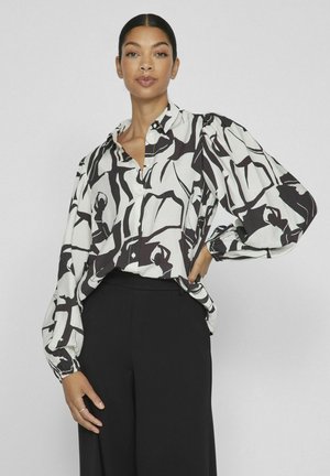 Blusa de patrón en blanco y negro con cuello, mangas largas sueltas y corte relajado, combinada con pantalones negros.