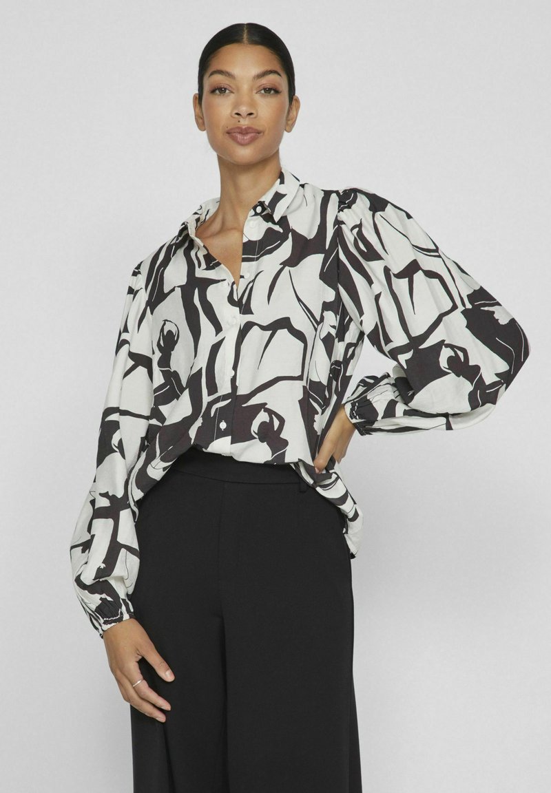 Blusa de patrón en blanco y negro con cuello, mangas largas sueltas y corte relajado, combinada con pantalones negros.