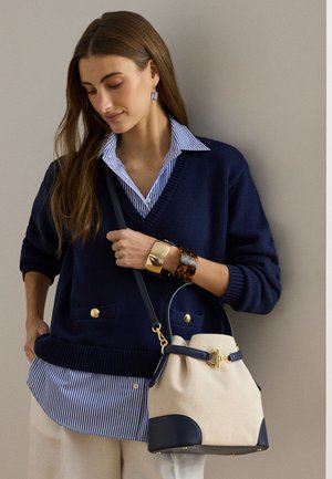 Γυναίκα με μπλε σκούρο πουλόβερ πάνω από μπλε ριγέ πουκάμισο, κρεμ παντελόνι, κρατάει μπεζ και μπλε σκούρο τσάντα με χρυσές λεπτομέρειες και φοράει βραχιόλια και σκουλαρίκια.