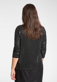 Czarna bluzka z długim rękawem o teksturowanym wzorze i delikatnym połysku, z dopasowanymi rękawami i prostym krojem, zestawiona z ciemnymi spodniami.