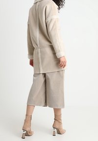 Chaqueta de imitación de piel de color beige forrada con pelaje, de corte relajado, con bolsillos laterales y detalle en la costura trasera, combinada con pantalones de pierna ancha de color beige y botines de tacón.