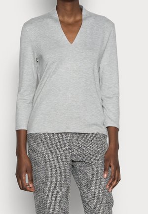 Person iført en lysegrå V-udskæring langærmet top og sort-hvide mønstrede bukser, stående mod en ensfarvet baggrund.