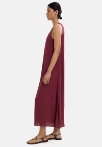 Bordowa, bezrękawnikowa sukienka maxi wykonana z lekkiego materiału, z luźnym krojem i bocznymi rozcięciami, zestawiona z płaskimi brązowymi sandałami.