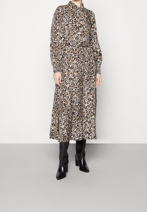 Kvinde iført en langærmet, knappet, leopardmønster midi kjole med taljebånd og sorte, hælede knæhøje støvler, stående på en grå gulv.