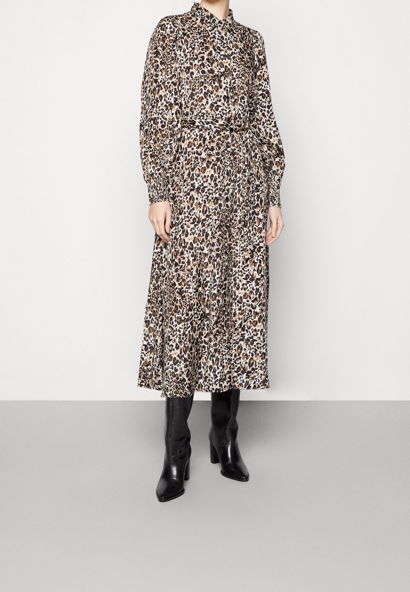Femme portant une robe midi à manches longues boutonnée, imprimé léopard, ceinture à la taille et bottes noires à talons montant jusqu'au genou, debout sur un sol gris.