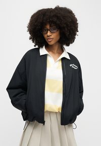 Γυναίκα με κατσαρά μαλλιά και γυαλιά, φορούν μαύρο μπουφάν με φερμουάρ πάνω από κίτρινο και λευκό ριγέ πουκάμισο με γιακά και μπεζ φούστα με πιέτες.