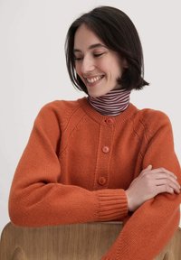 Orange Strickjacke mit Rundhalsausschnitt und vier vorderen Knöpfen. Mit gerippten Bündchen und strukturierten Mustern an den Ärmeln.