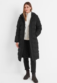 Long black puffer coat with a hood, featuring a zip front and quilted design. Worn over a white top and paired with black pants and boots.