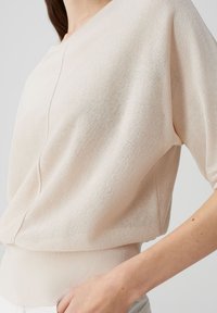 Helles beige Kurzarmoberteil mit strukturiertem Finish, das über einen runden Ausschnitt und einen gerippten Saum verfügt. Lockere Passform mit dezenten Nahtdetails.