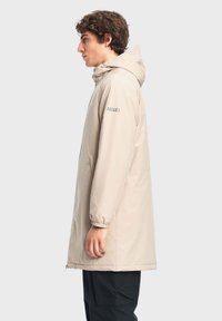 Bež vodootporna parka s kapuljačom, elastičnim manžetama i dva bočna džepa. Sadrži logo na lijevom rukavu. Glatka tekstura i srednje dugački dizajn.
