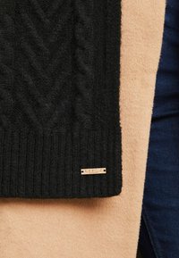 Sort strikket tørklæde med et kabelmønster, ribbede kanter og et guldhardware mærke. Sat mod en blød beige stofbaggrund.