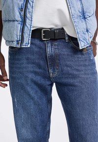 Jeans in denim di un blu scuro con una texture sfumata. Abbinati a un gilet trapuntato azzurro chiaro e a una cintura in pelle nera con fibbia argentata.