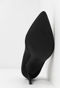 Chaussure à talon stiletto noir à bout pointu, inclinée contre un mur blanc, montrant la semelle inférieure et le talon fin.