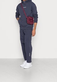 Man i navy Adidas hoodie och joggers med vita sneakers och en burgunderfärgad crossbody-väska, stående mot en ljus, plain bakgrund.