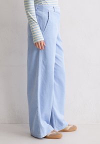 Hellblaue Cord-Hose mit weitem Bein und vertikalen Rillen, ausgestattet mit seitlichen Taschen und einem lockeren Sitz, kombiniert mit beigen und braunen Schuhen.