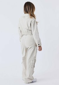 Chaqueta recortada en beige claro con cinturilla elástica, combinada con pantalones cargo a juego que tienen bolsillos laterales y perneras ajustadas. Tejido texturizado.