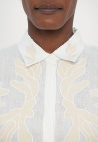 White linen blouse featuring a button-up front and a collar, adorned with cream-colored abstract embroidery patterns.