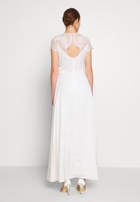 Robe blanche avec une superposition de dentelle aux épaules, un dos ouvert et une jupe fluide, assortie à des chaussures à talons hauts en satin.