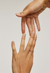 Anillo de plata con grabado de "uno" en la banda, reposando sobre una mano con uñas naturales, mientras otra mano se acerca a él. Textura de piel suave.