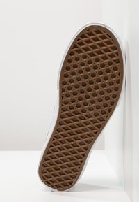 Vit sneaker med en texturerad, brun gummisula som har ett diamantmönster. Överdelen har inga synliga snören eller hårdvarudetaljer.