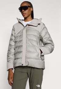 Giacca puffer grigia chiara con una superficie liscia e leggermente lucida, dotata di zip frontale, cappuccio e pattern a griglia sul corpo.