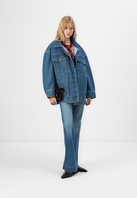 Woman wearing oversized blue denim jacket, matching flared jeans, burgundy sweater, black pointed shoes, and holding a small black clutch.