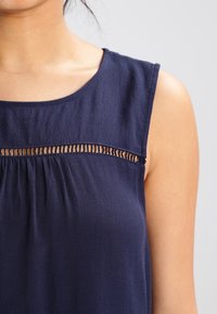 Ciemnoniebieska bluzka bez rękawów z okrągłym dekoltem, z poziomą przezroczystą wstawką i marszczoną fakturą poniżej piersi.