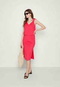 Vestuário rosa sem mangas com decote em V e fenda lateral, combinado com sandálias pretas e óculos de sol grandes. Modelo segura uma bolsa de palha.