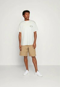 T-shirt de algodão cinza claro com mangas curtas e um pequeno design impresso no peito, combinado com calções bege e sapatilhas brancas.