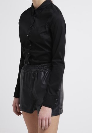 Person wearing a fitted black button-up long sleeve shirt and black high-waisted elastic leather shorts, standing against a neutral background.